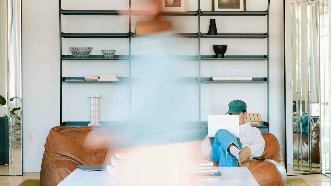 A person sits on a brown beanbag chair using a laptop, while another person walks by in front of them, appearing blurred from motion. The background features minimalist shelves with books and decorative objects.